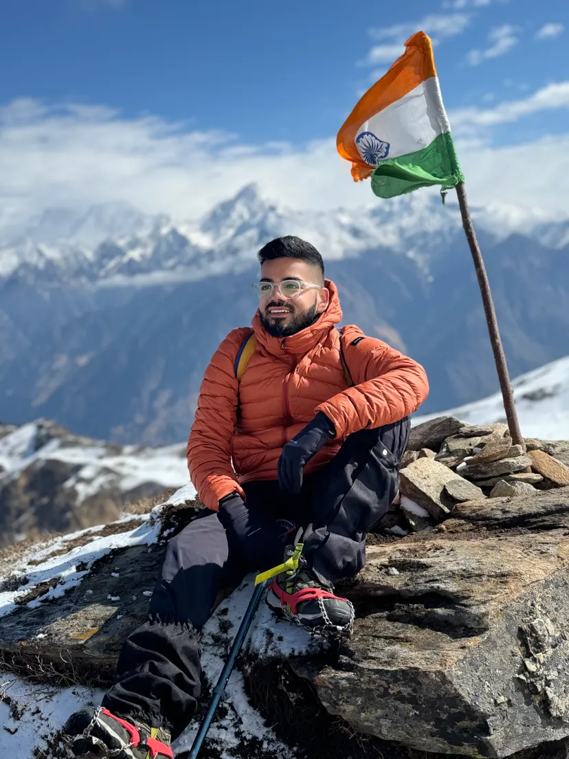 Kush in an orange down jacket on a snowy mountain summit beside the Indian flag