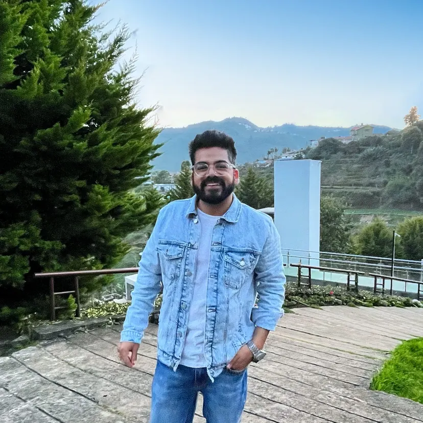 Kush in a denim jacket on a wooden deck, with forested mountains behind him