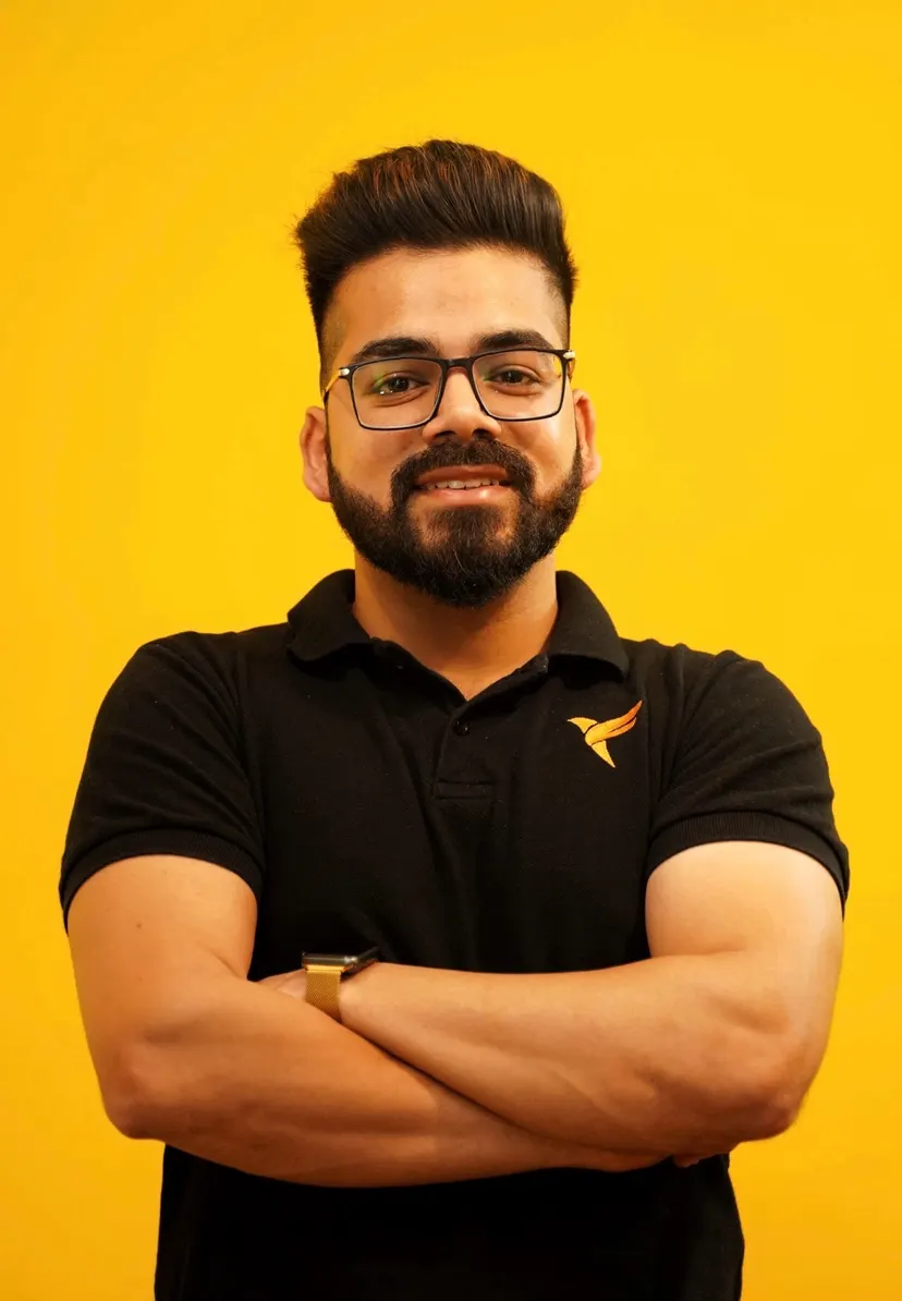Kush with his arms crossed in a black polo, against a yellow wall